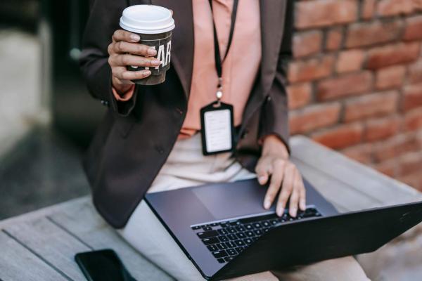 Woman wearing an ID badge on a lanyard holds a coffee in one hand and types on a laptop with the other.