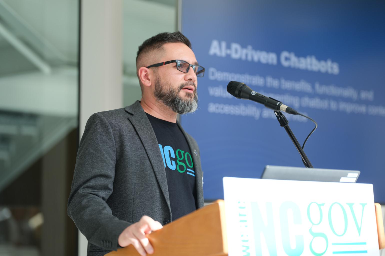 A man wearing a We Are NC Gov shirt speaks at a podium. A screen showing a slide on AI-Driven Chatbots is behind him.