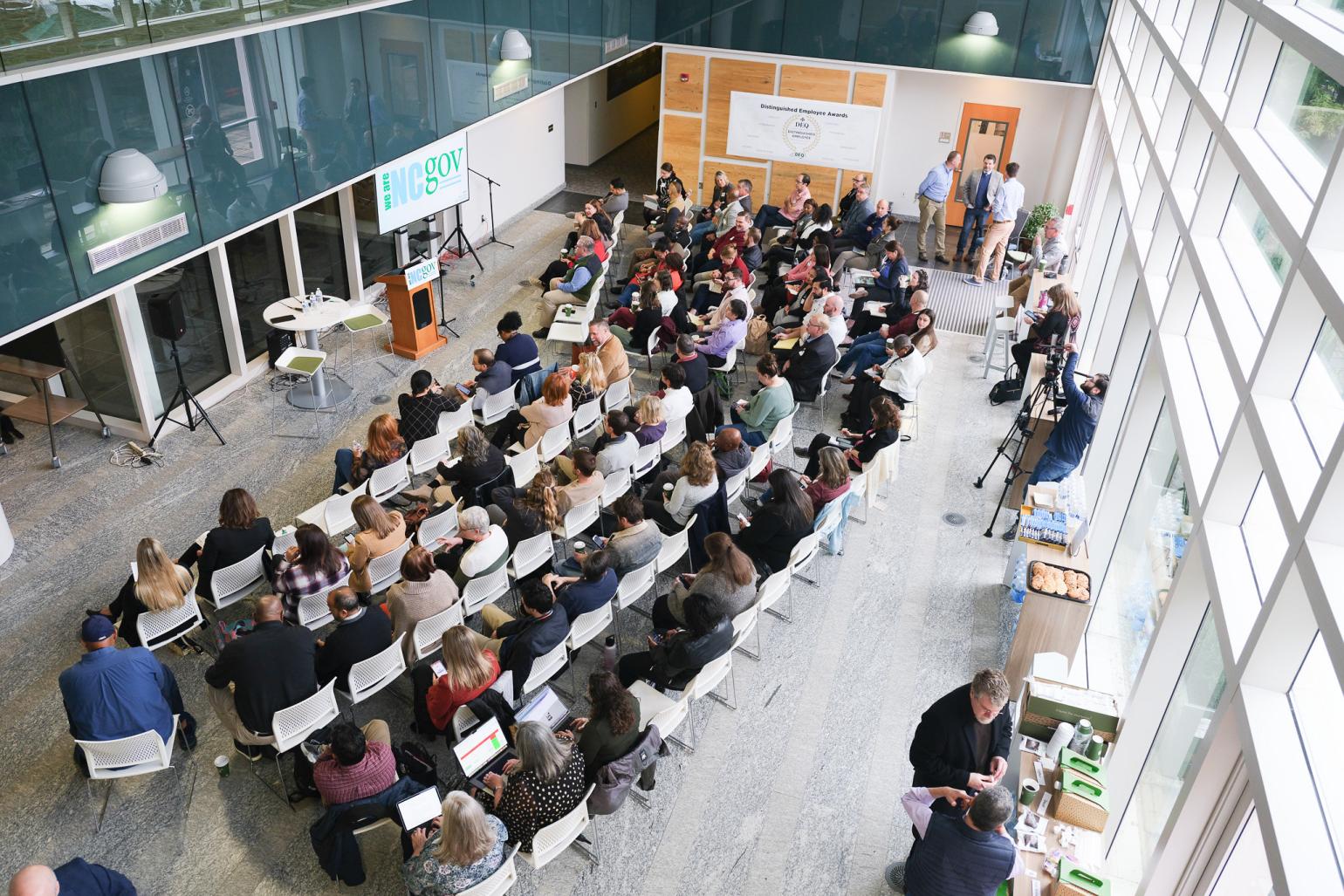 Lines of chairs are filled in an auditorium with a wall of glass windows.