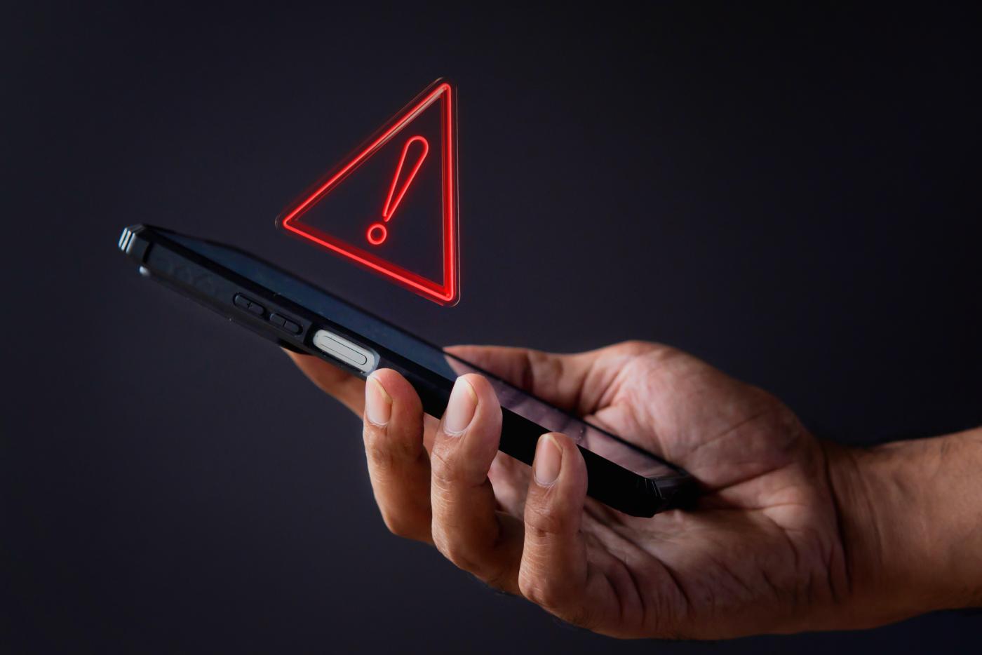 The image shows a hand holding a cell phone with a red warning symbol above it