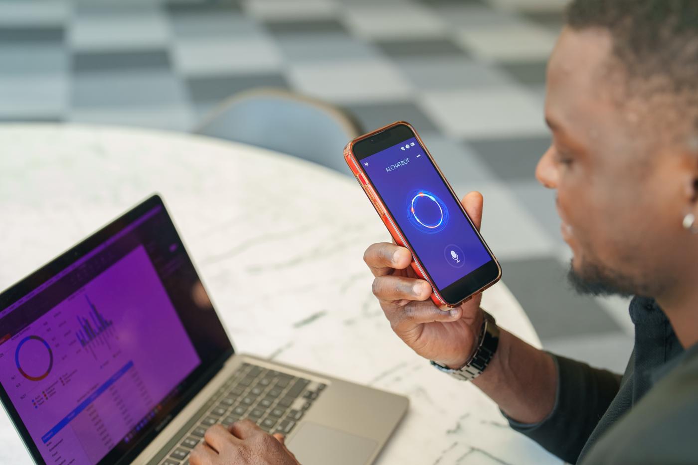 A person is sitting at a table using a laptop displaying a data analytics screen with charts and graphs. They are holding a smartphone with a screen showing an AI chatbot. 