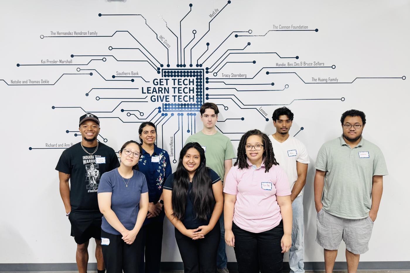 Group photo of interns standing in front of a wall at the Kramden Institute that says "Get Tech, Learn Tech, Give Tech"