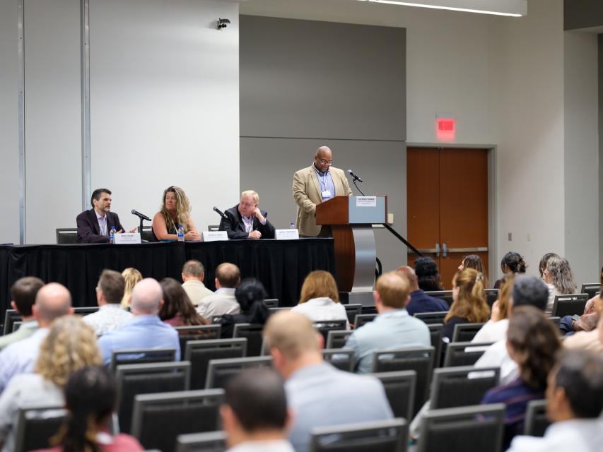 Innovation Director Raymond Thomas speaks from a podium with other panelists seated at a table next to him at the N.C. Digital Government Summit