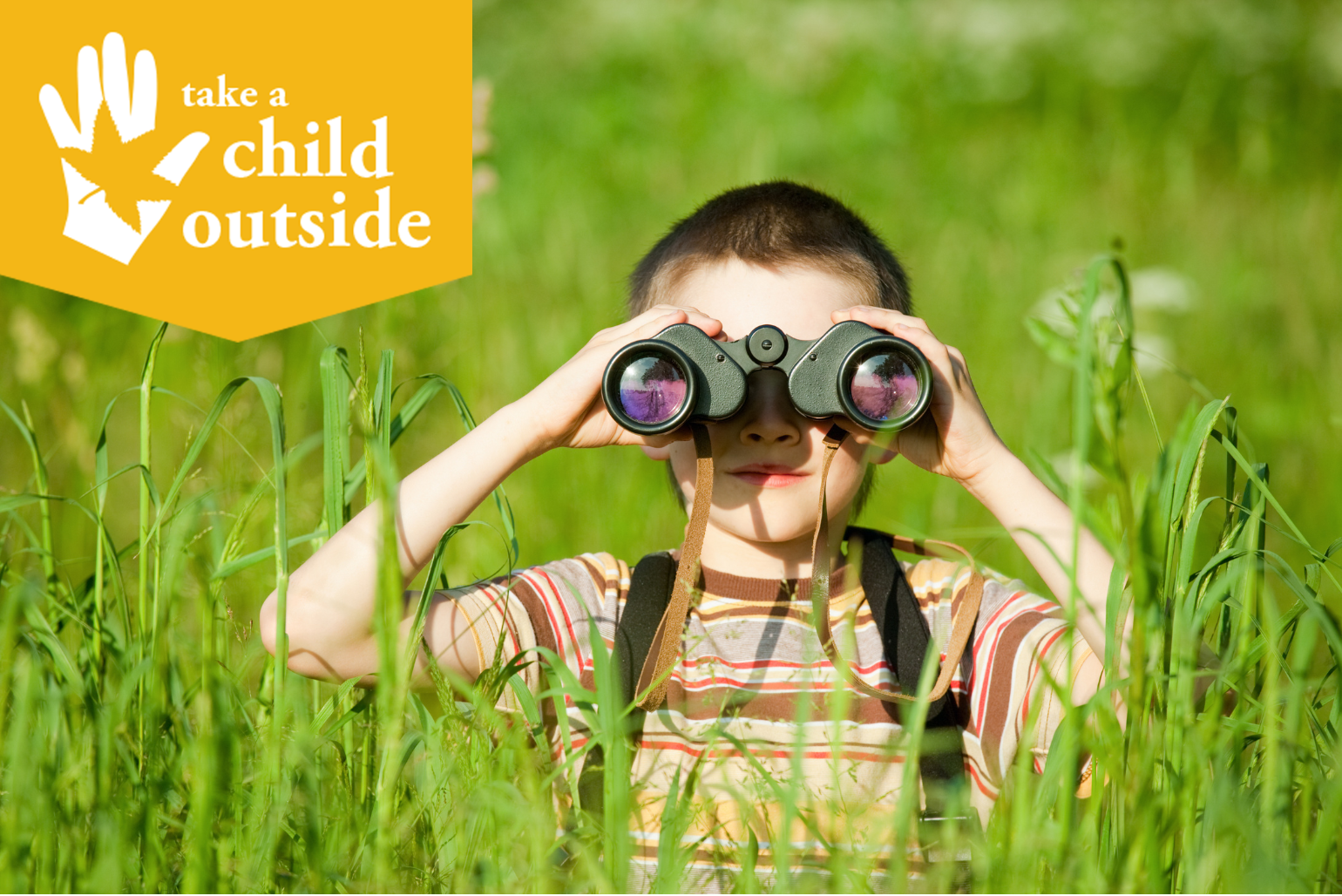 A boy sits in tall grass and looks out with binoculars.