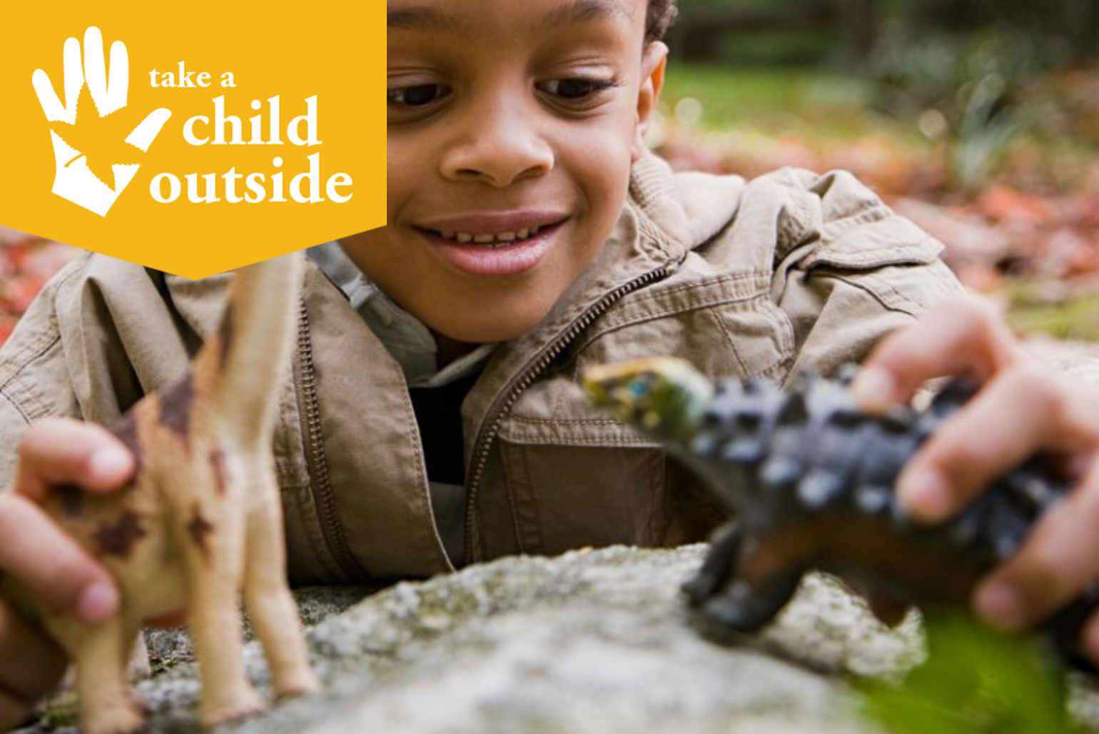 A little boy plays with dinosaurs outside on a rock.