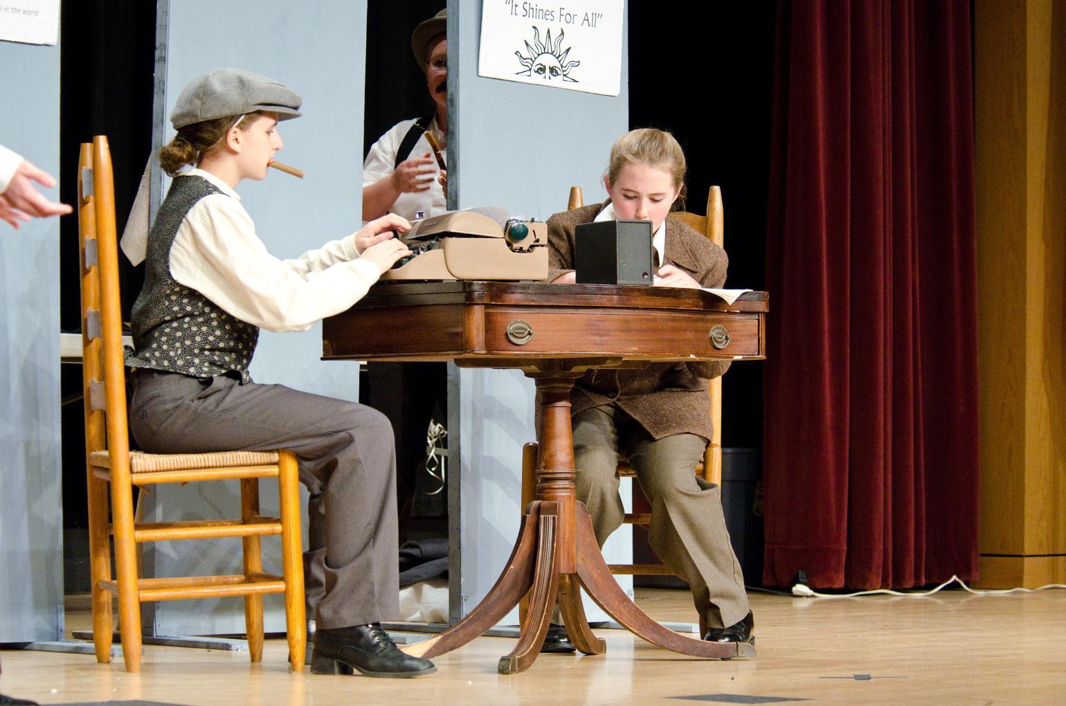 Two students in historic costumes sit at a table on stage during their History Day performance.