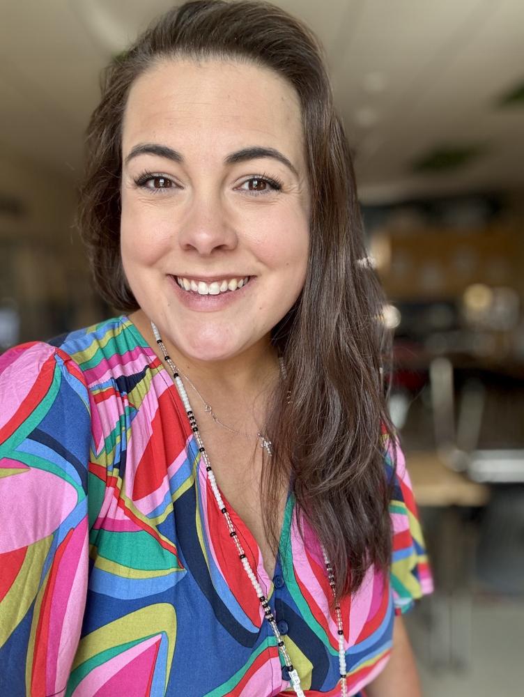a woman with long brown hair wears a colorful dress and smiles at the camera