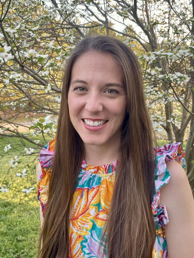 a woman with long brown hair wears a colorful dress and smiles at the camera