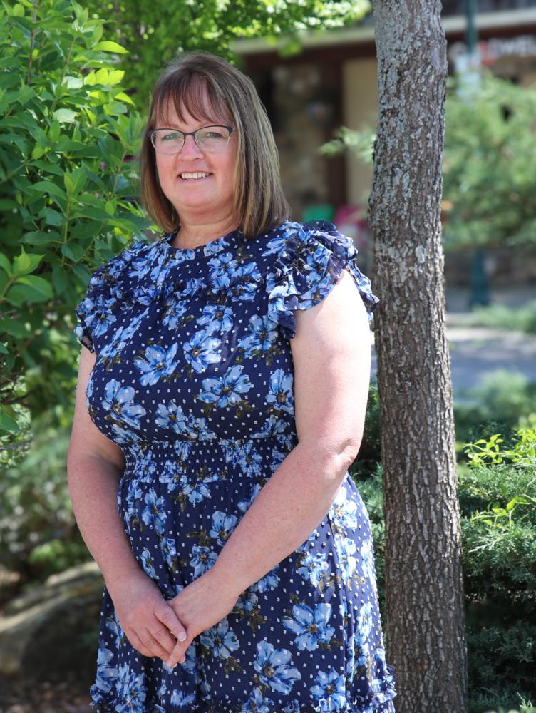 a woman with chin length brown hair and wearing a blue dress stands in front of a tree while smiles at the camera