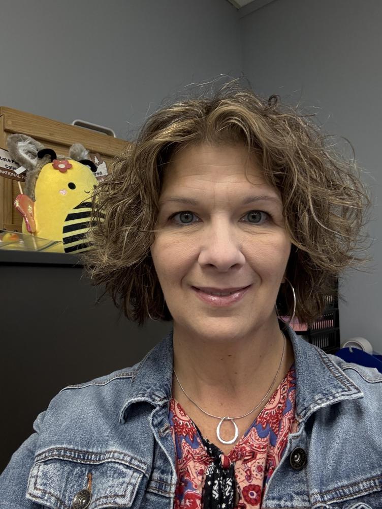 a woman with shoulder length curly brown wears a jean jacket and smiles at the camera