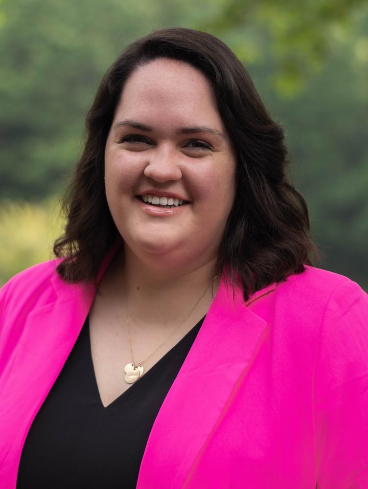 a woman with shoulder length brown hair wears a bright pink jacket and smiles at the camera