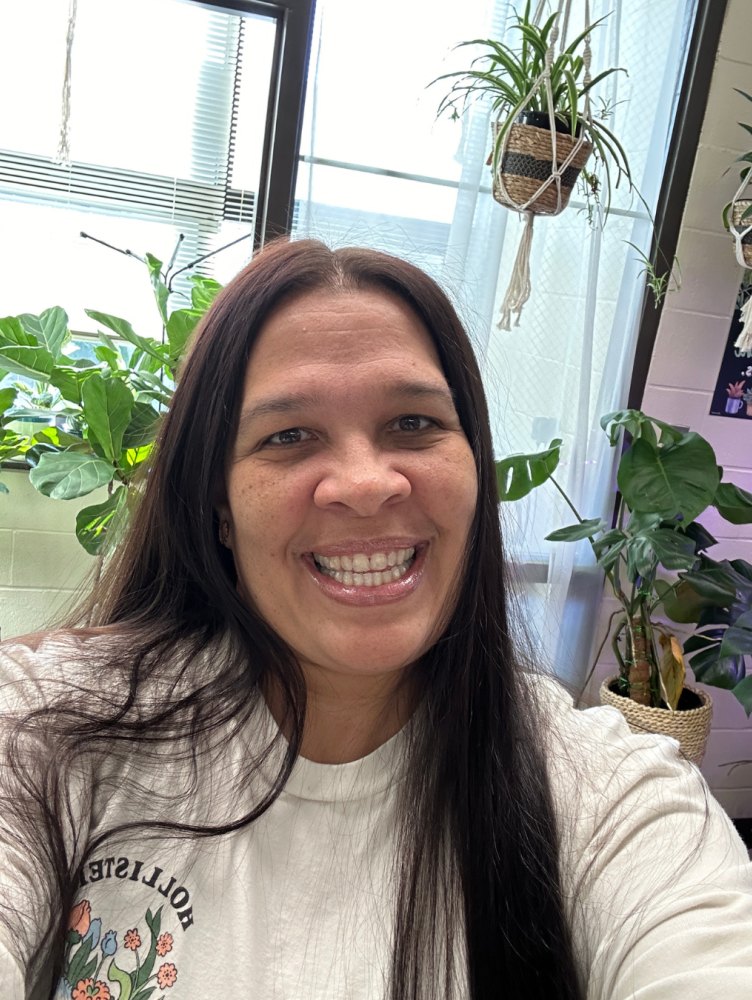 a woman with long dark hair smiles in front of plants in a window