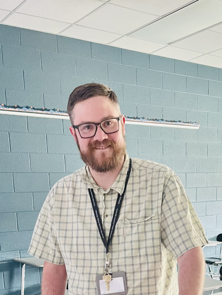 a white man with glasses and a plaid shirt stands in a classroom and smiles at the camera