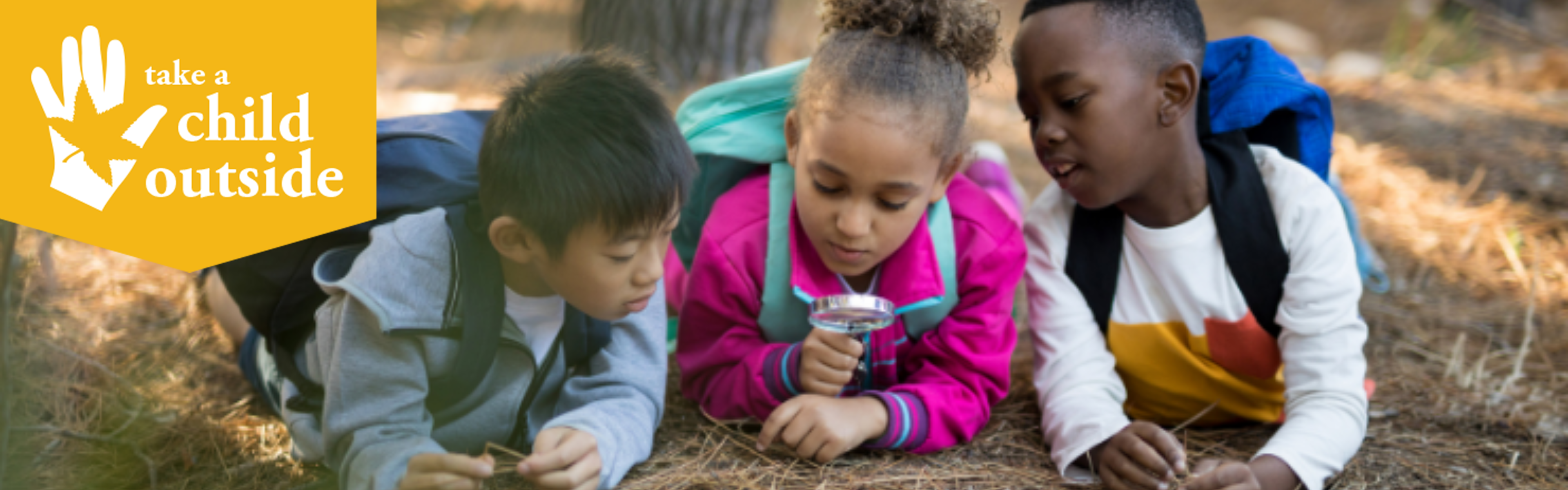 Three kids lay on the ground in a pine forest and look at the ground with a magnify glass.