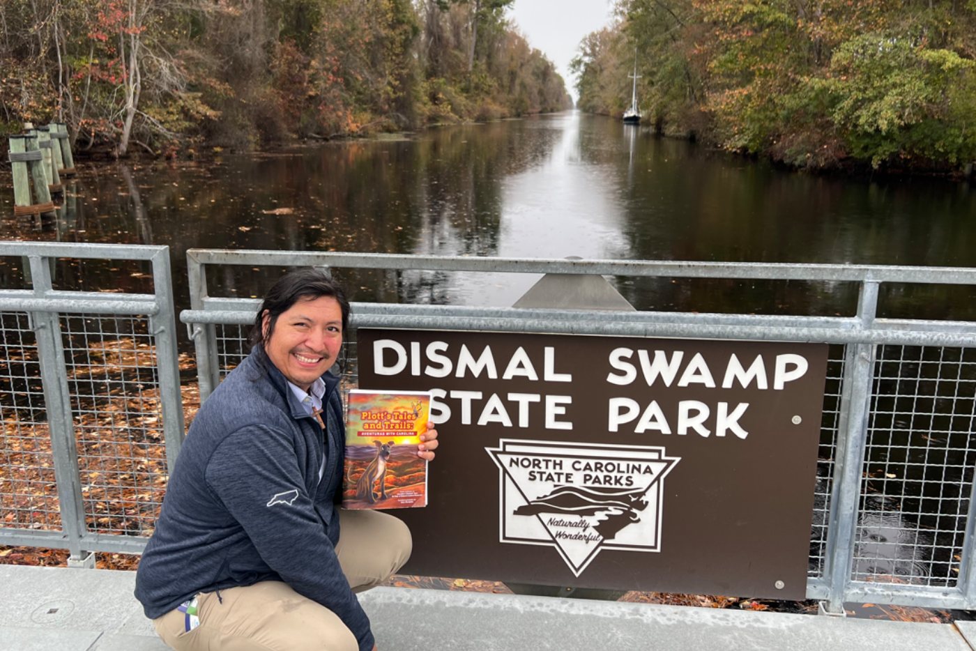 Hugo holds a copy of the Plott's Tales and Trails book as he squats in front of a canal at Dismal Swamp State Park.