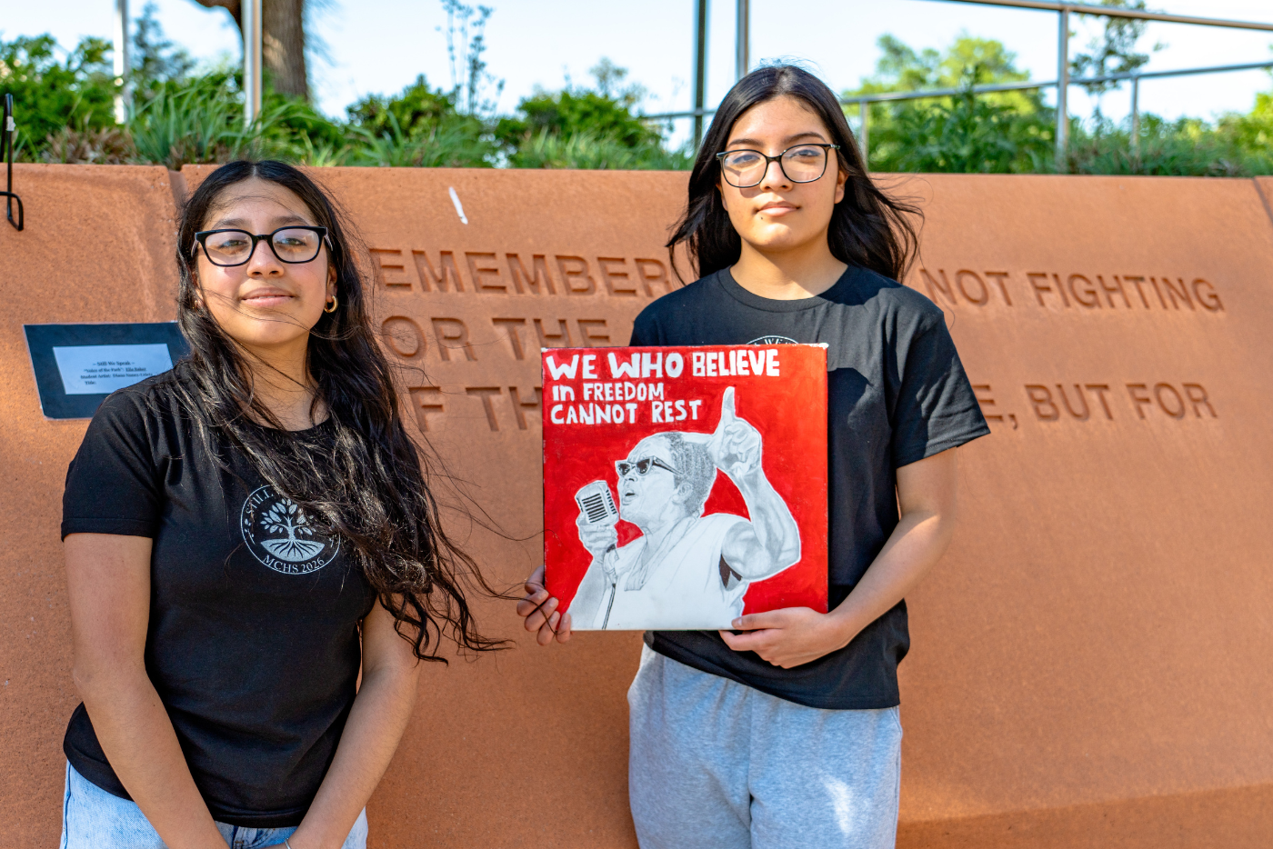 Two high school students stand in Freedom Park holding artwork that reads "We who believe in freedom cannot rest."  