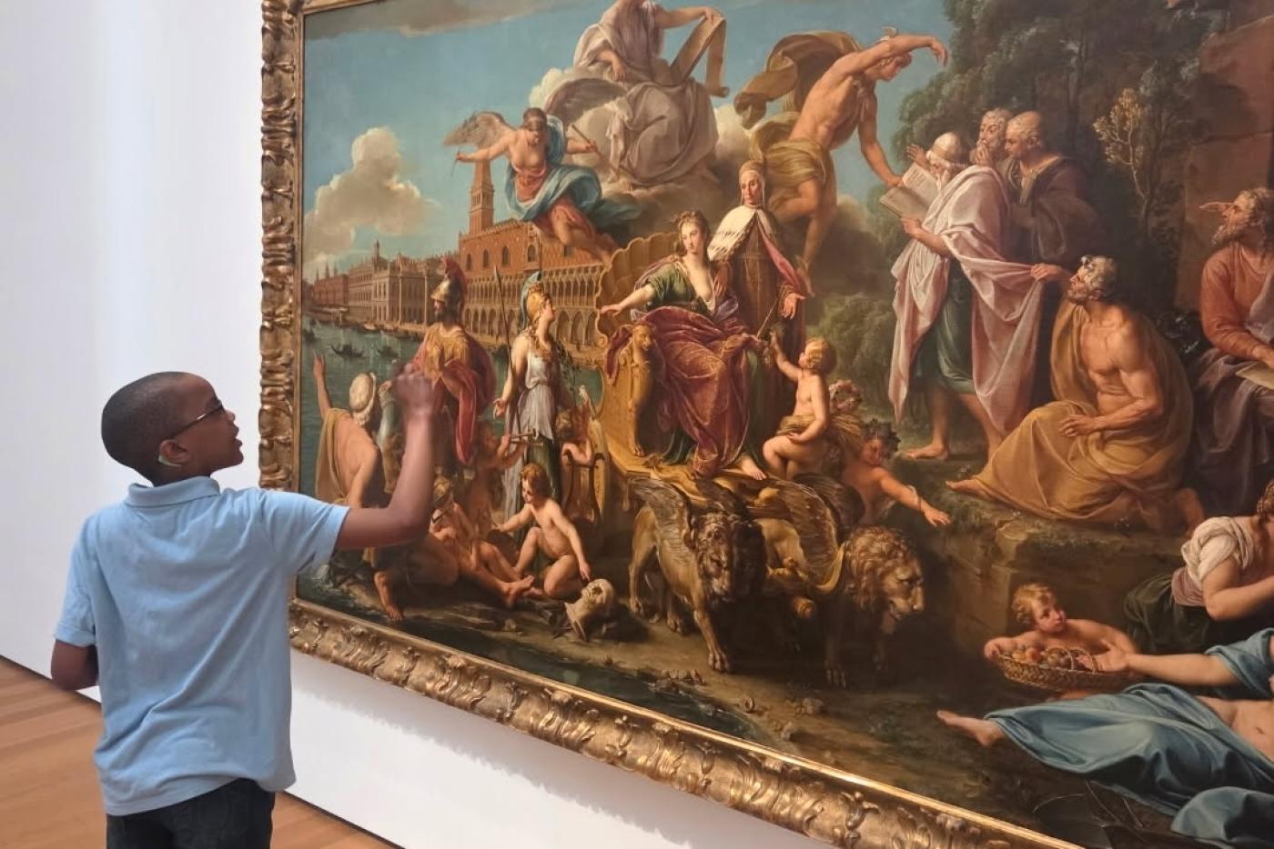 a young African American student stands in front of a piece of Renaissance style art, looking at it and pointing to different elements. 
