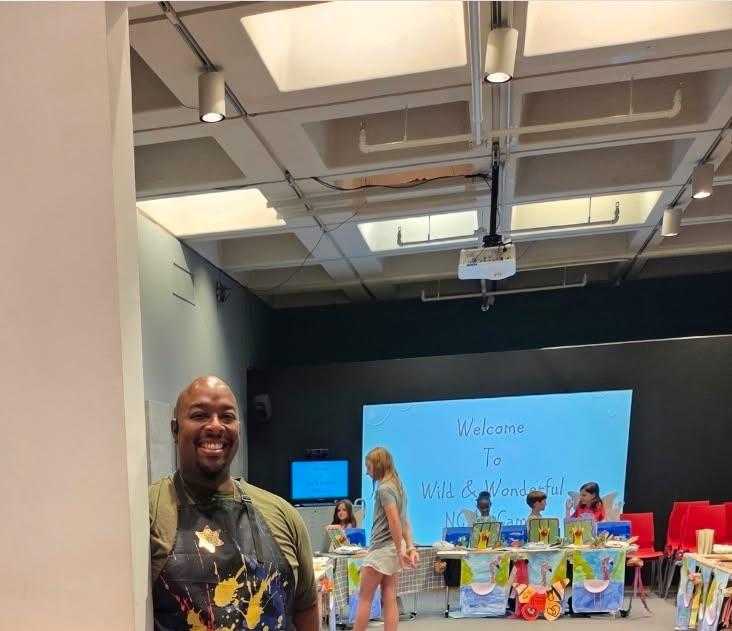 A smiling African American Man stands at the doorway to a room filled with children working on art projects