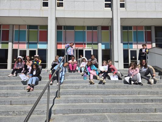 A group of students smile while sitting on the front steps of the State Library and State Archives of North Carolina.