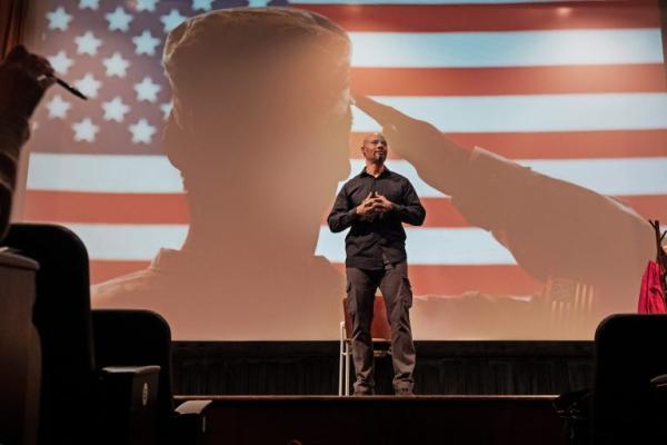 Dr. Sonny Kelly stands in front of a screen showing an American Soldier saluting the American Flag.