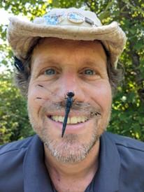 A male state parks ranger smiles at the camera with a dragon fly on his nose.