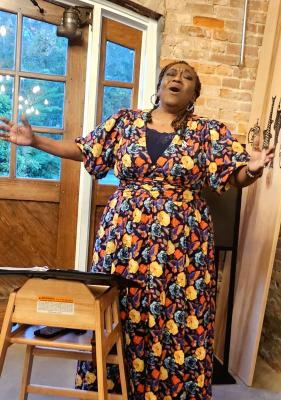 An African American woman in a floral dress sings with arms outstretched
