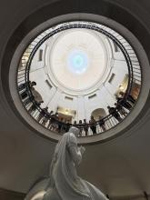 A ground up view of a marble George Washington Statue under a Capitol Rotunda ceiling. Students looking down at camera
