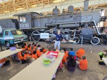 Students wearing orange shirts sit in front of an instructor and a steam locomotive
