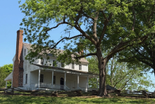 A two-story and double chimney white colonial house sits behind a tree and split rail fence
