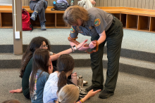 A park ranger holds an orange snake for seated students. One student reaches out to pet the snake.