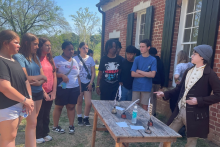An 18th century living history interpreter points to a wooden table and writing quills to teach students about quill writing