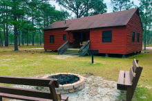 A historic red cabin with a fire pit and two benches are set within a wooded forest