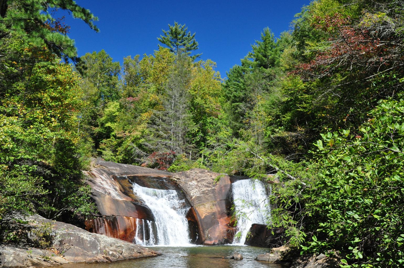 two waterfalls cascade over brown rock into a pool with green trees surrounding them at Gorges State Park