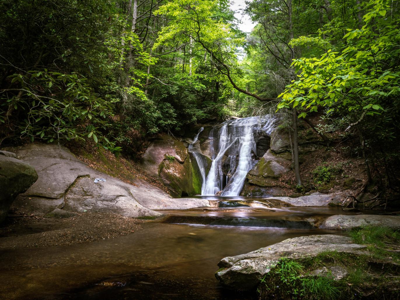 a waterfall flows over mossy rock into a pool surrounded by boulders and lush green forest at Stone Mountain State Park