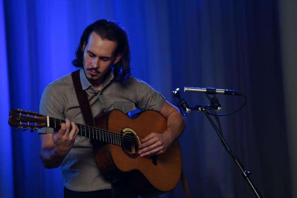 Lakota John plays a guitar next to a microphone in front of a blue curtain