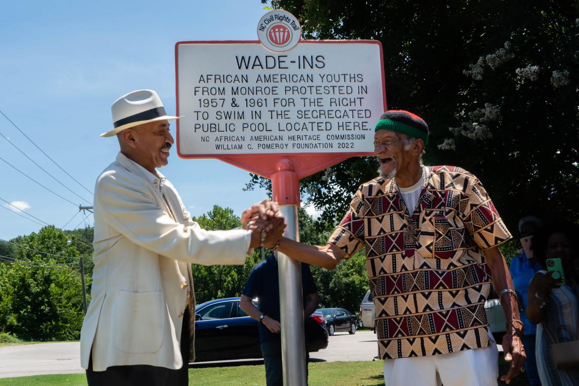 Two elder Black men clasp hands in front of a Civil Rights Trail Marker