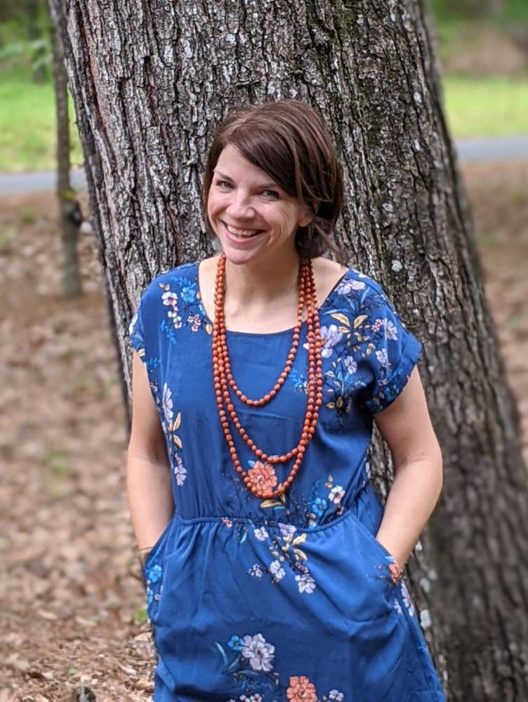 a white woman with chin length brown hair wears a blue dress and leans against a tree