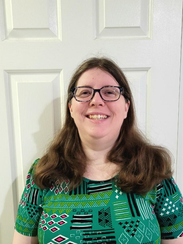 a white woman with glasses and shoulder length brown hair wears a green patterned dress
