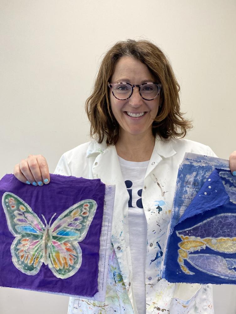 a white woman in a white lab coat holds up two pieces of art 
