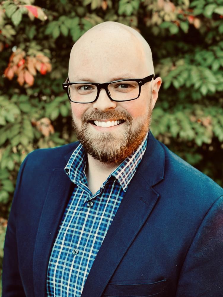 a man with glasses and a blue suit jacket and plaid shirt smile at the camera