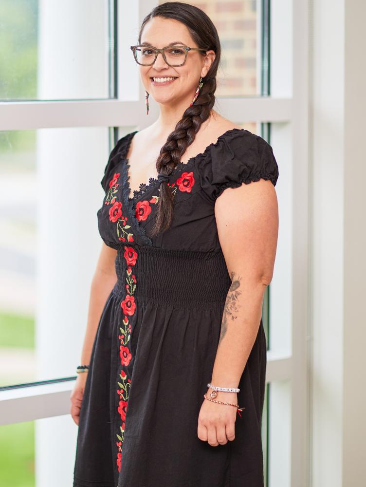 a woman with a long black braid stands dressed in a black and red floral dress in front of a window