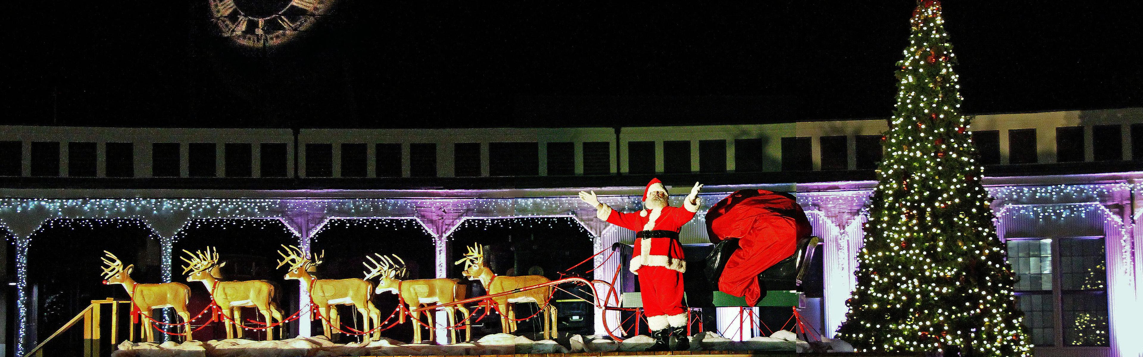 Santa Clause gestures towards the camera as he stands between a christmas tree and a sleigh with five plastic reindeer.
