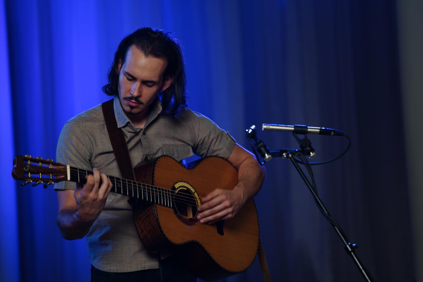 Lakota John plays a guitar next to a microphone in front of a blue curtain