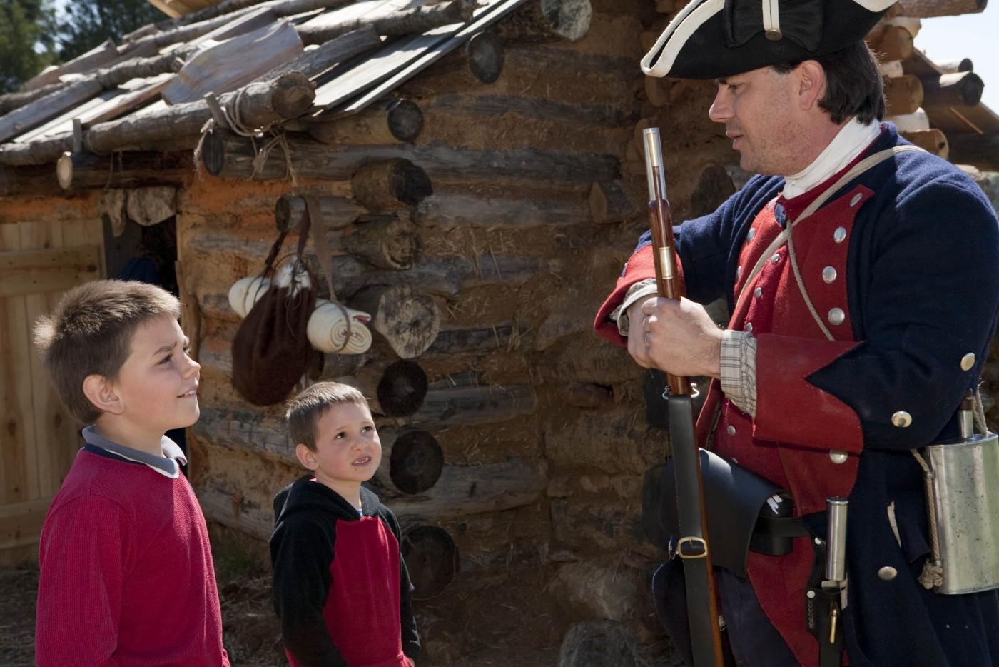 A historical interpreter dressed as a Revolutionary era soldier speaking with two children.