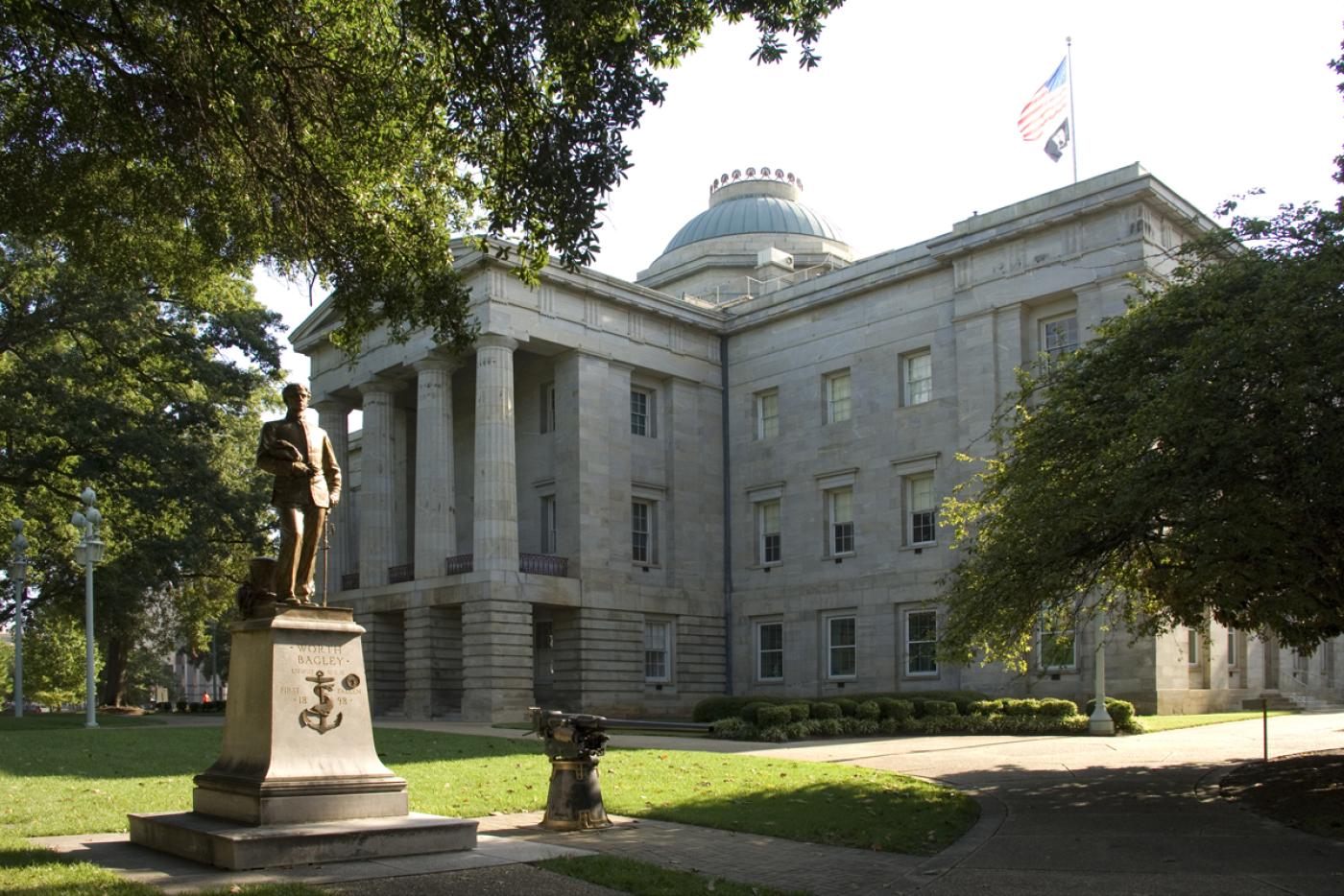A side view of the North Carolina State Capitol building. A statue can be seen in front of the building.