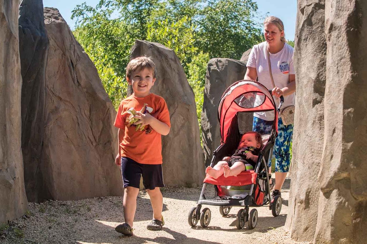 Mother pushing a stroller behind a toddler walking between large rocks on each side