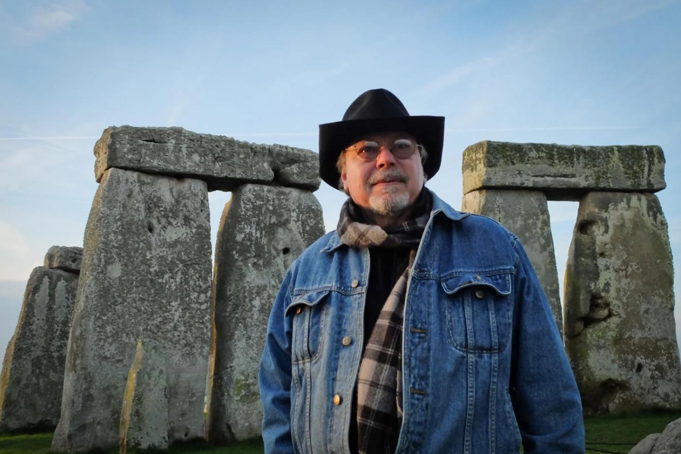 A man in a jean jacket and cowboy hat standing in front of Stonehenge.