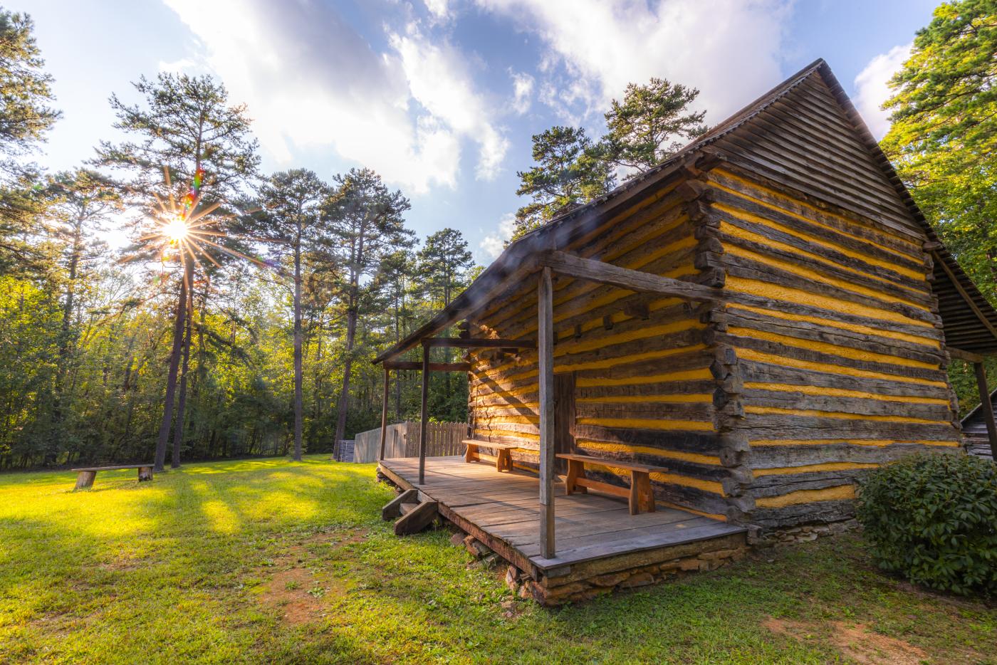 The Allen House, a log dwelling at Alamance Battleground, sits on green grass with tall pine trees surrounding it.