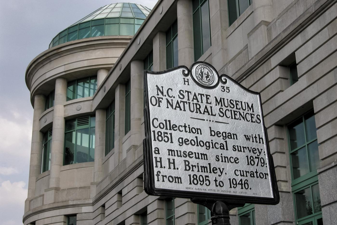 A historical marker featuring information about the Library of NC State Museum of Natural Sciences with a grey stone building with a glass dome behind it 