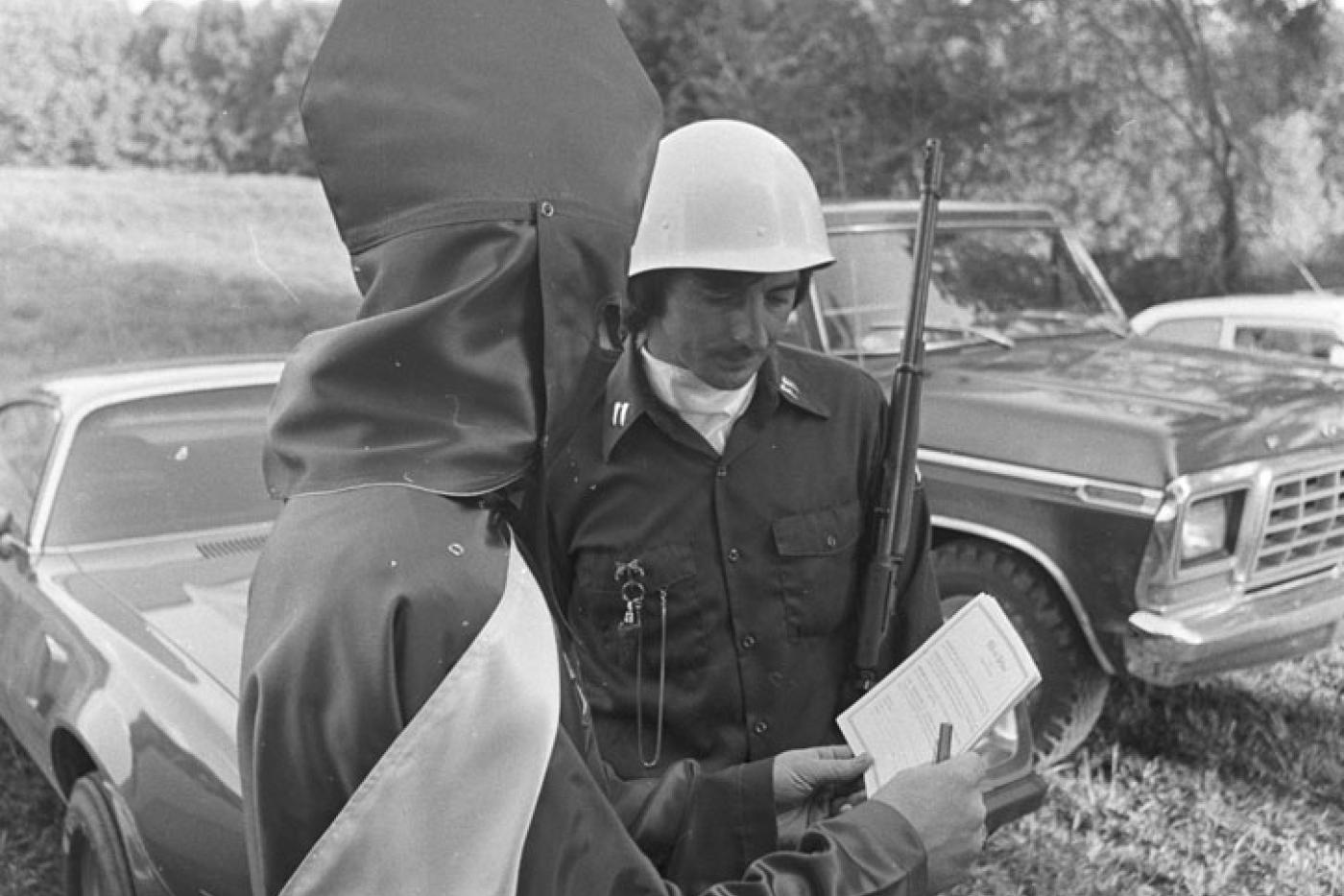 A police officer holding a rifle, speaking with a Ku Klux Klan member holding up a piece of paper.